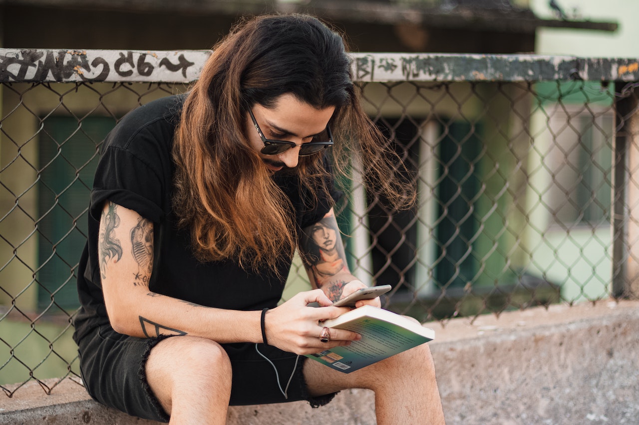 A man with long hair sitting on a low wall reading a book