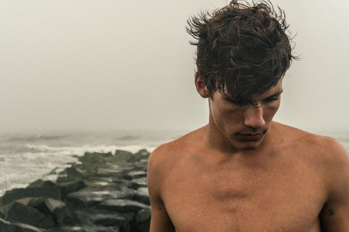 Man poses shirtless with coastline rocks in the background