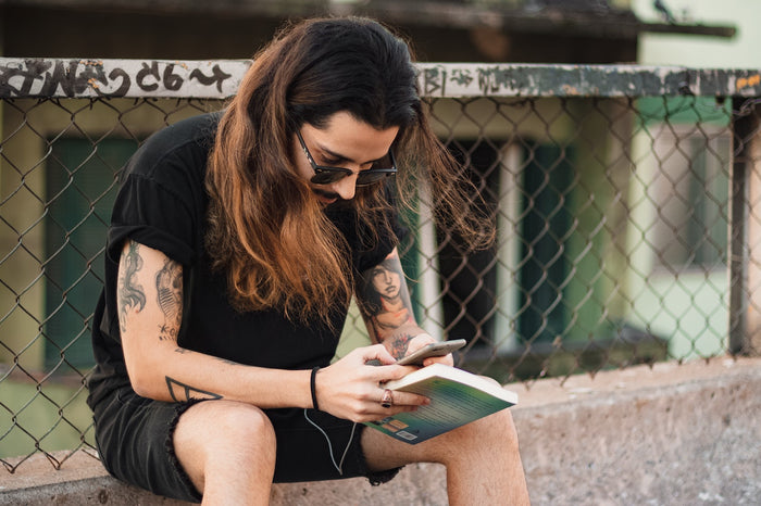 A man with long hair sitting on a low wall reading a book
